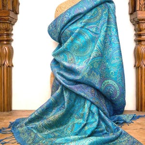 May include: A rectangular shawl featuring a vibrant turquoise and blue base with an intricate, multi-colored paisley pattern in shades of gold, purple, and green. The fabric has a subtle sheen and is finished with matching turquoise fringe at the ends.