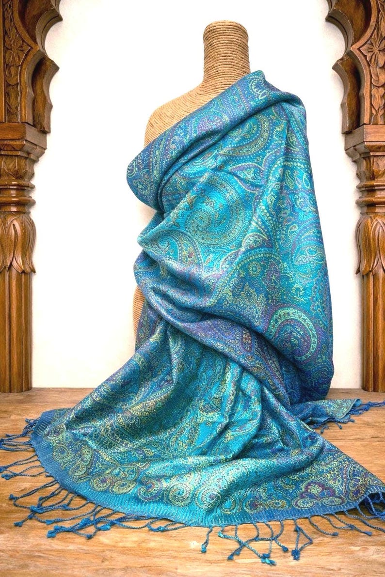 May include: A rectangular shawl featuring a vibrant turquoise and blue base with an intricate, multi-colored paisley pattern in shades of gold, purple, and green. The fabric has a subtle sheen and is finished with matching turquoise fringe at the ends.