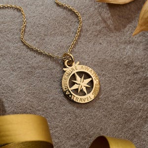 Op de afbeelding: Een goudkleurige kompas hanger ketting met de woorden "Adventure Awaits Travel" gegraveerd rond de kompasroos. De ketting ligt op een grijze ondergrond met gouden lint en gedroogde bladeren.