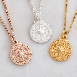 Op de afbeelding: Drie kompas kettinghangers in roségoud, zilver en goud. Elke ketting heeft een ronde hanger met een kompasroosontwerp en de woorden "Adventure Awaits Travel Safe" gegraveerd. Gepresenteerd op een wit marmeren oppervlak.