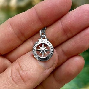 Op de afbeelding: Een zilverkleurige hangerketting met een kompasontwerp. De hanger heeft een cirkelvormig frame met de woorden "Adventure Awaits Travel Safe" rond een stervormige kompasroos. De ketting wordt in een hand gehouden.