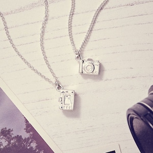 May include: Two silver camera pendant necklaces on a white wooden surface. One pendant is a classic camera design, the other is a vintage camera design. The necklaces are displayed with a photograph and a camera lens.