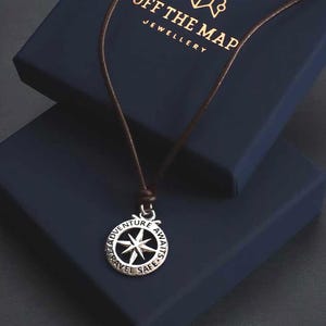 Op de afbeelding: Een zilveren kompas ketting met een bruin koord. De hanger heeft de woorden "Adventure Awaits, Travel Safe" rond het kompasroos ontwerp. De ketting wordt getoond op een donkerblauwe sieradendoos met "Off The Map Jewellery".
