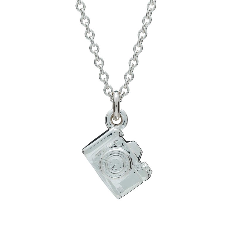 May include: A silver-colored necklace featuring a small camera pendant. The pendant is a detailed miniature replica of a camera, with visible lens and body details. The chain is a delicate, linked design.
