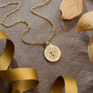 Op de afbeelding: Een goudkleurige ketting met een kompas hanger. De hanger heeft de woorden "Adventure Awaits, Travel Safe" gegraveerd rond een kompasroos ontwerp. De ketting ligt op een grijze ondergrond met gouden lint en gedroogde bladeren.
