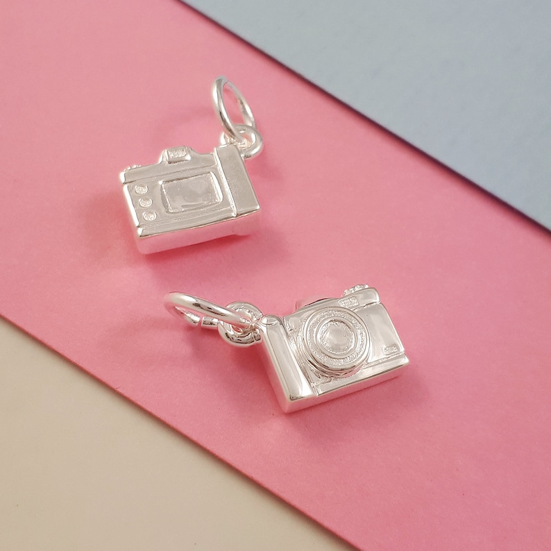 May include: Two silver-colored camera charms with detailed designs. The charms have a loop at the top for attaching to a necklace or bracelet. The background is a combination of pink and blue.