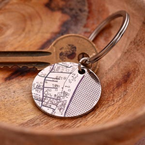 May include: A circular silver keychain with a detailed map design. The map shows a mix of straight lines and geometric shapes, with a section of wavy lines. A key and key ring are attached.