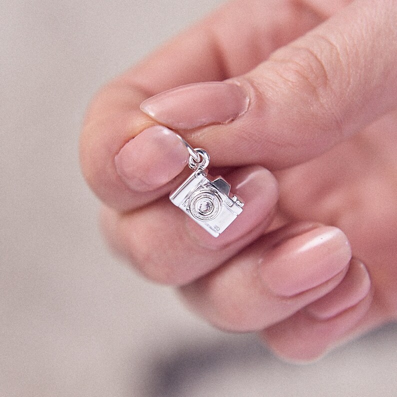 May include: A small, silver-colored camera charm with intricate details, including a lens and flash. The charm is suspended from a small loop, suggesting it's designed for a necklace or bracelet. The camera charm is held in a hand.
