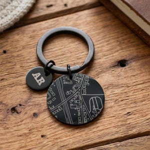 Könnte beinhalten: Ein Schlüsselanhänger aus mattschwarzem Metall mit einem runden Anhänger, der mit einem weißen Straßenkartenmotiv graviert ist, sowie einem kleineren runden Anhänger mit den Buchstaben AR in Serifenschrift an einem stabilen Metallring.