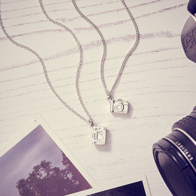 May include: Two silver camera pendant necklaces on a white wooden surface. One pendant is a classic camera design, the other is a vintage camera design. The necklaces are displayed with a photograph and a camera lens.