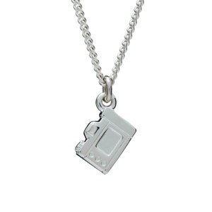 May include: A silver-colored necklace featuring a small camera pendant. The pendant is a detailed miniature replica of a camera, with visible buttons and a lens. The chain is a delicate, linked design, suitable for everyday wear.