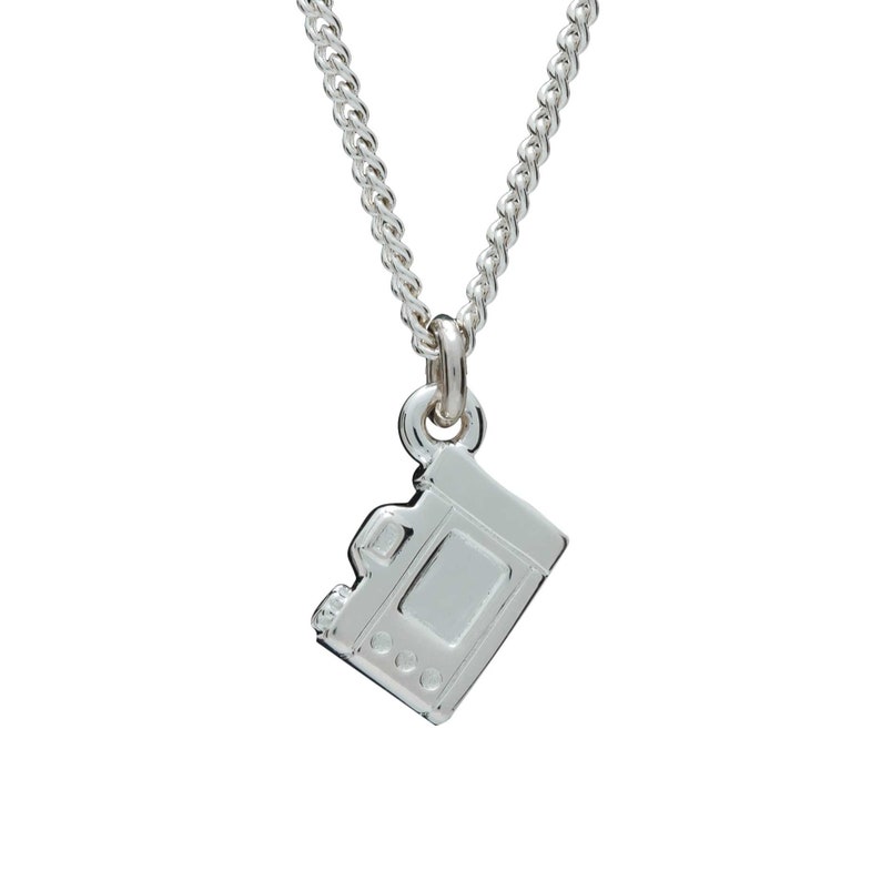 May include: A silver-colored necklace featuring a small camera pendant. The pendant is a detailed miniature replica of a camera, with visible buttons and a lens. The chain is a delicate, linked design, suitable for everyday wear.