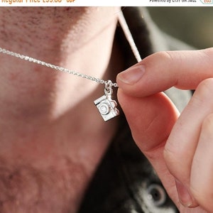 May include: A silver necklace featuring a small camera pendant. The camera charm has intricate details, including a lens and buttons. The necklace is held by a person, showcasing the delicate chain and the pendant's design.