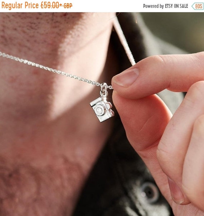 May include: A silver necklace featuring a small camera pendant. The camera charm has intricate details, including a lens and buttons. The necklace is held by a person, showcasing the delicate chain and the pendant's design.