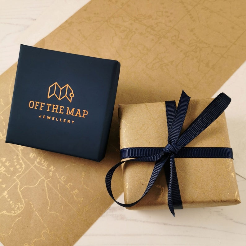 May include: A navy blue jewelry box with the text "OFF THE MAP JEWELLERY" and a gold map logo, next to a small, wrapped gift tied with a navy blue ribbon. The background features a map design.