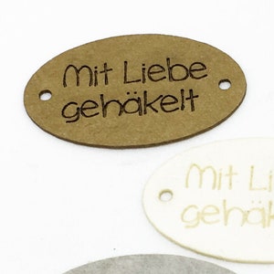 Peut inclure: Quatre étiquettes ovales avec le texte "Mit Liebe gehäkelt" en différentes couleurs : marron, blanc, gris et noir.
