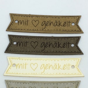 Può includere: Cinque etichette di carta marrone, beige e nero con il testo "mit ♥ gehäkelt" in bianco.
