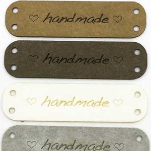 Può includere: Cinque etichette in pelle con la parola "fatto a mano" e un cuore su ciascuna. Le etichette sono di colore marrone, marrone scuro, bianco, grigio e nero.