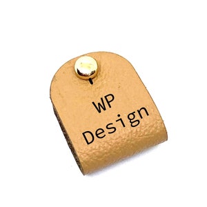 Op de afbeelding: Een lichtbruine leren snoerorganizer met een gouden metalen knoop en de tekst "WP Design" erop gedrukt.