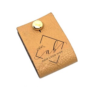 Op de afbeelding: Een bruine leren stempel met een gouden metalen knop. De stempel heeft een vierkant frame met een bloemmotief en de tekst "ab" en "DEIN LOGO HIER".