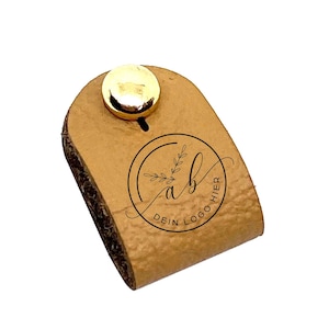 Op de afbeelding: Een lichtbruine leren band met een gouden metalen knoop en een zwart rond logo met de tekst "Dein Logo Hier" met een gestileerde "ab" en een klein blaadjesontwerp.