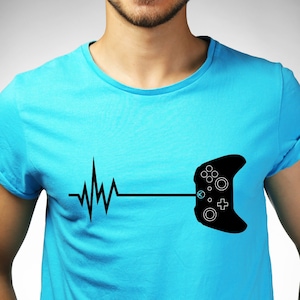 Puede incluir: Un hombre con una camiseta azul claro con un gráfico negro de un mando de videojuego y una línea de latido del corazón.