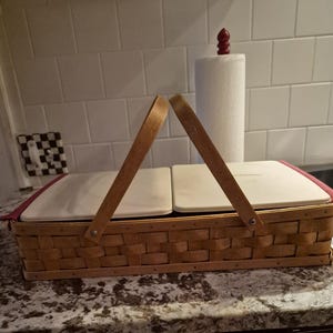 May include: A rectangular woven wood basket featuring two hinged wooden handles and two white rectangular lids. The basket has a maroon fabric liner visible along the top edges, and the woven structure displays a natural light brown wood finish.