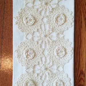 May include: A white kitchen towel with crocheted lace trim and six crocheted flower designs with pearl accents.