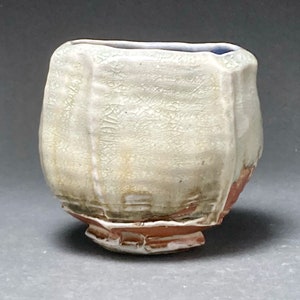 May include: A white ceramic teacup with a textured surface and a slightly uneven rim. The cup has a light brown glaze on the bottom and a blue glaze on the inside rim.