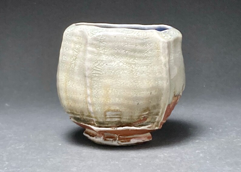 May include: A white ceramic teacup with a textured surface and a slightly uneven rim. The cup has a light brown glaze on the bottom and a blue glaze on the inside rim.