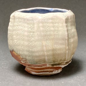 May include: A small, white ceramic cup with a blue glaze on the inside. The cup has a textured surface and a slightly irregular shape.