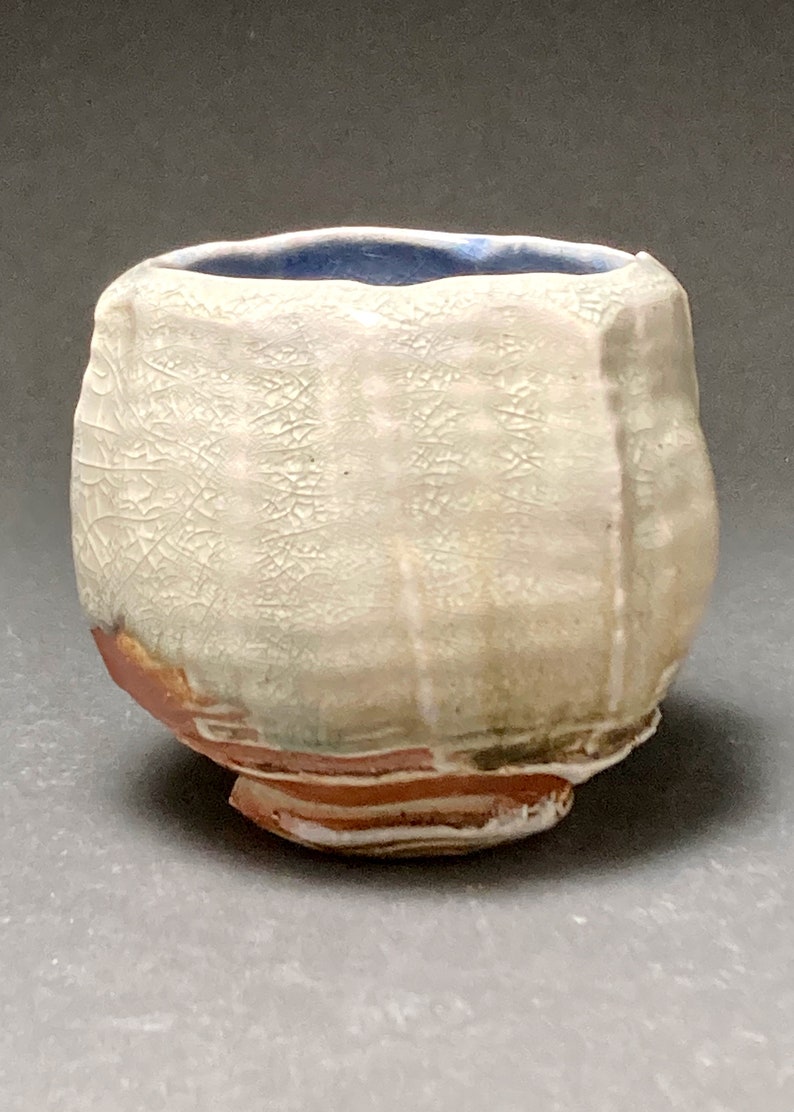 May include: A small, white ceramic cup with a blue glaze on the inside. The cup has a textured surface and a slightly irregular shape.