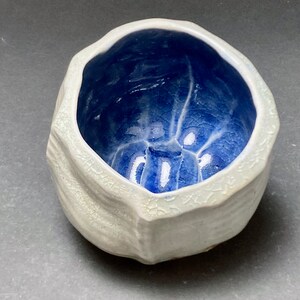 May include: A small, white ceramic bowl with a textured exterior and a blue glazed interior. The bowl has an irregular, organic shape.