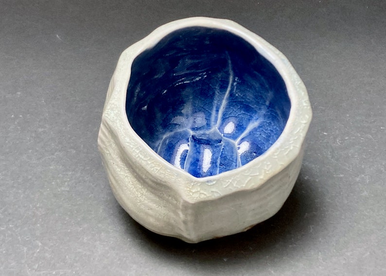 May include: A small, white ceramic bowl with a textured exterior and a blue glazed interior. The bowl has an irregular, organic shape.