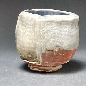 May include: A small, white ceramic cup with a textured surface and a brown rim. The cup has a unique, irregular shape and a slightly uneven base.