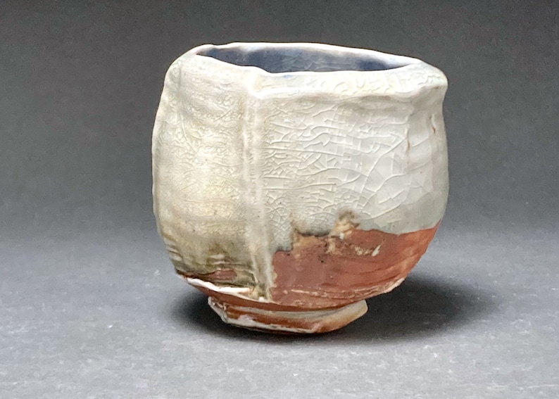 May include: A small, white ceramic cup with a textured surface and a brown rim. The cup has a unique, irregular shape and a slightly uneven base.