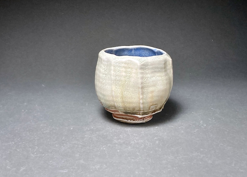May include: A small, white ceramic cup with a blue glaze inside and a brown glaze on the bottom. The cup has a textured surface and a rounded shape.