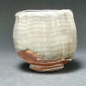 May include: A white ceramic cup with a crackled glaze and a brown and green base. The cup has a rounded shape and a small foot.
