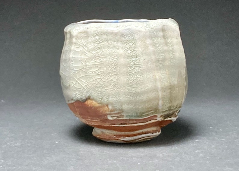 May include: A white ceramic cup with a crackled glaze and a brown and green base. The cup has a rounded shape and a small foot.
