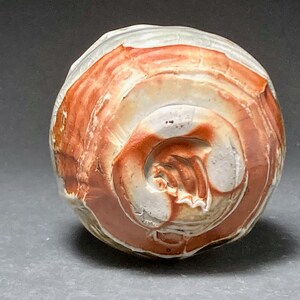 May include: A small, round ceramic pot with a white and red glaze. The pot has a textured surface and a unique swirl pattern.