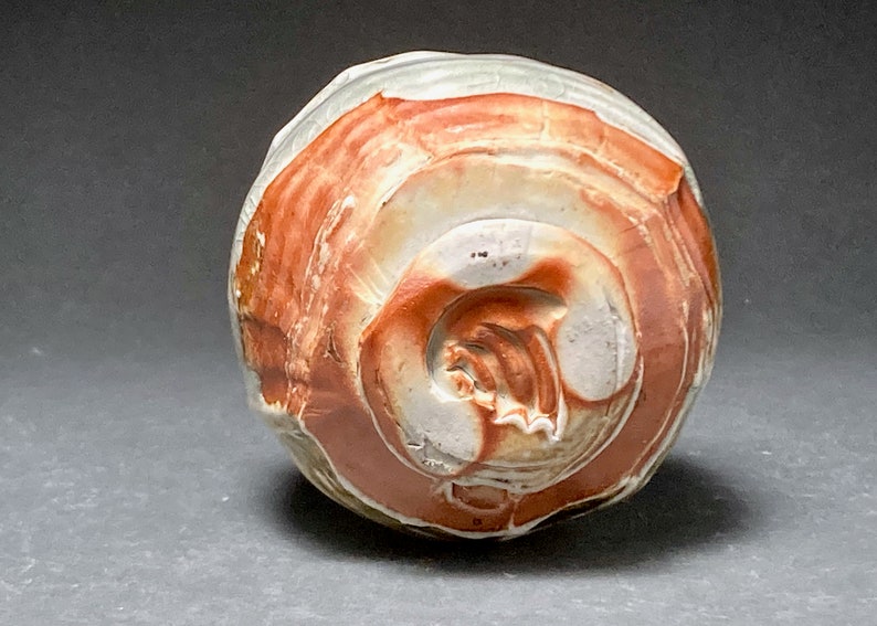 May include: A small, round ceramic pot with a white and red glaze. The pot has a textured surface and a unique swirl pattern.