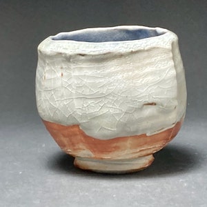 May include: A small, white ceramic cup with a crackled glaze and a light blue rim. The cup has a rounded base and a slightly flared lip. The bottom of the cup is a reddish-brown color.
