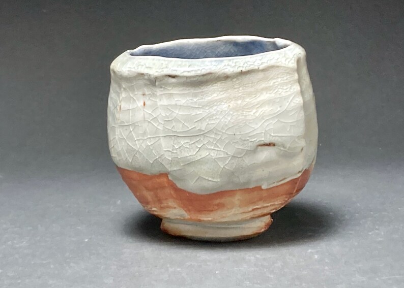 May include: A small, white ceramic cup with a crackled glaze and a light blue rim. The cup has a rounded base and a slightly flared lip. The bottom of the cup is a reddish-brown color.