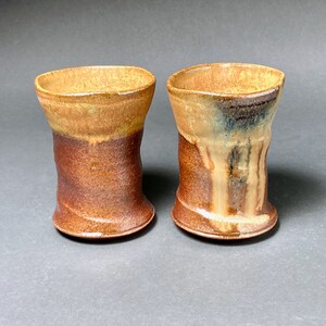 May include: Two ceramic mugs with a brown and yellow glaze. The mugs have a unique, slightly uneven shape.