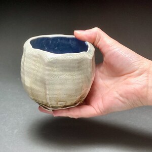 May include: A small, white ceramic cup with a blue glaze inside. The cup has a textured, uneven surface and a slightly rounded shape.