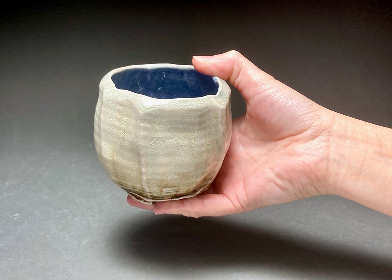 May include: A small, white ceramic cup with a blue glaze inside. The cup has a textured, uneven surface and a slightly rounded shape.