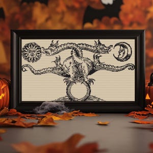 May include: Black framed artwork featuring a detailed black and white illustration of a mythical creature with a sun and moon. The artwork is displayed against a backdrop of autumn leaves and pumpkins, evoking a Halloween theme.