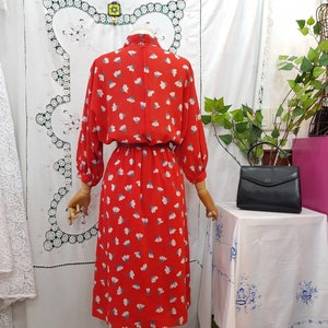 May include: A vintage red dress with a floral pattern of white and blue flowers. The dress has long sleeves, a high neckline, and a fitted waist. A black handbag sits on a table next to the dress.