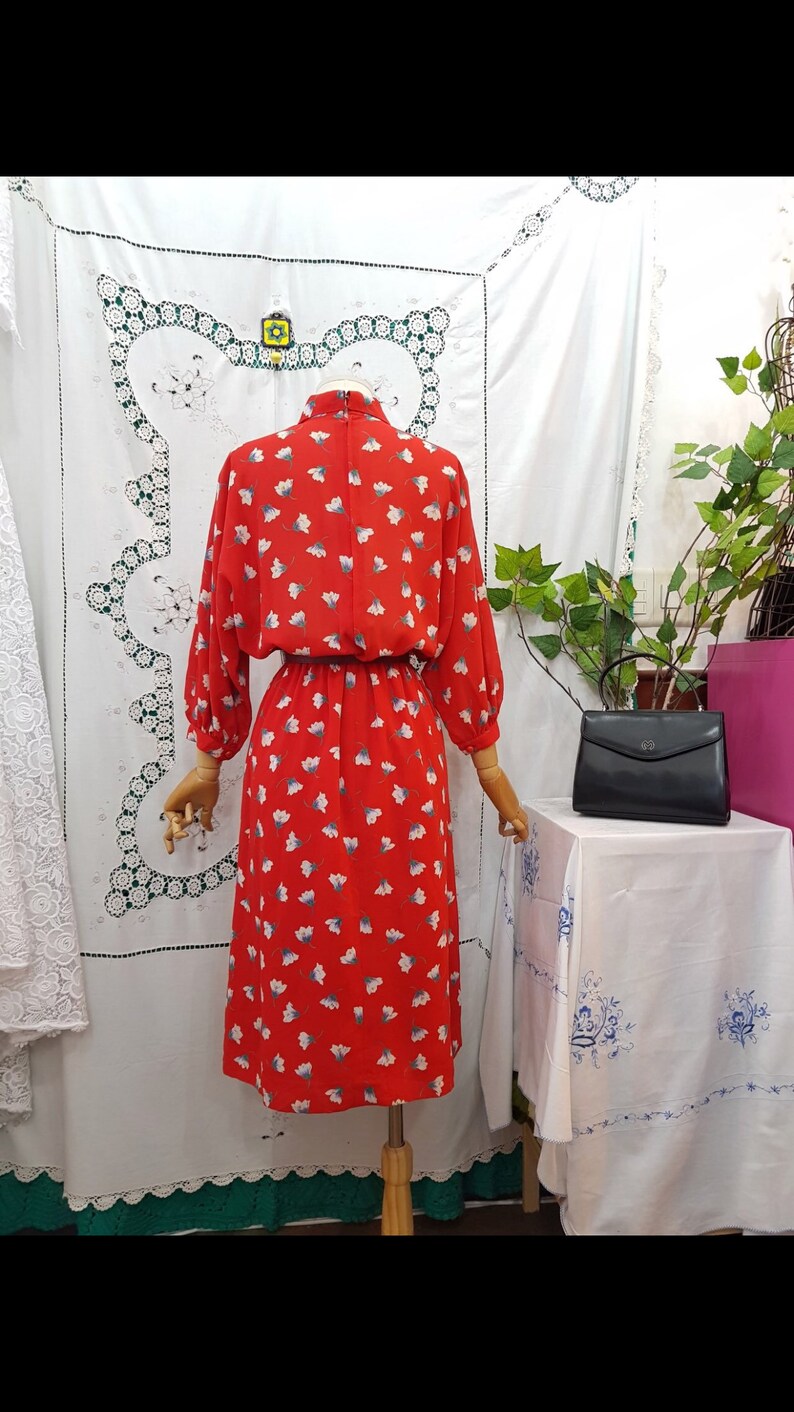 May include: A vintage red dress with a floral pattern of white and blue flowers. The dress has long sleeves, a high neckline, and a fitted waist. A black handbag sits on a table next to the dress.