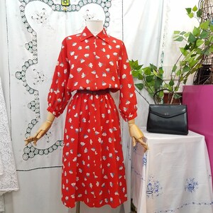 May include: A vintage red dress with a white floral pattern, long sleeves, and a collared neckline. The dress is displayed on a mannequin. A black handbag sits on a table nearby. The dress has a belt at the waist.